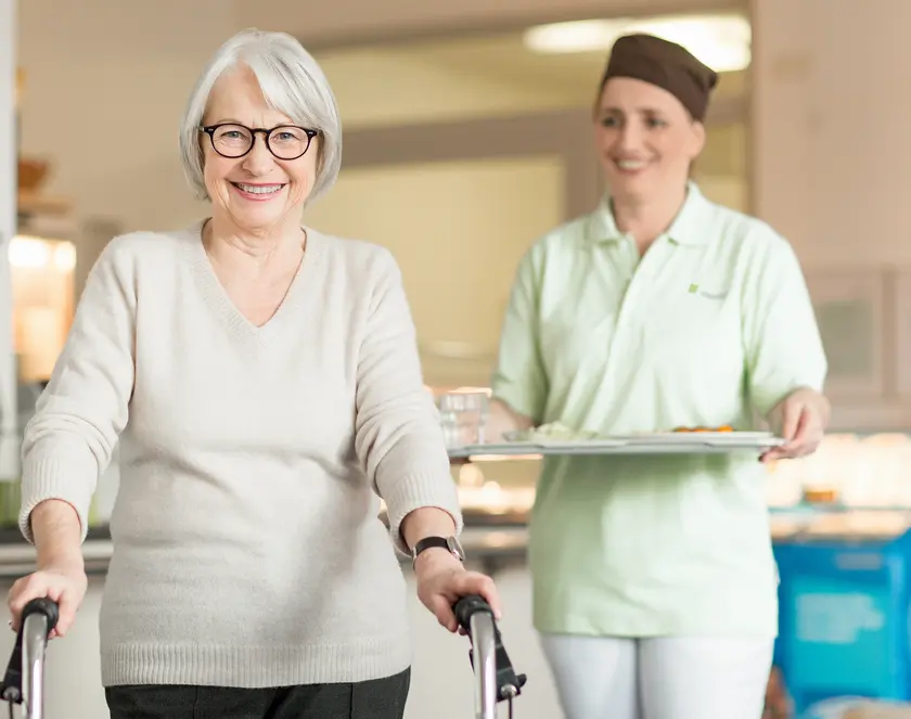 WISAG Mitarbeitende trägt Speisentablet für Seniorin mit Rollator