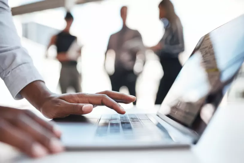 Person nutzt einen Laptop während im Hintergrund weitere Personen stehen